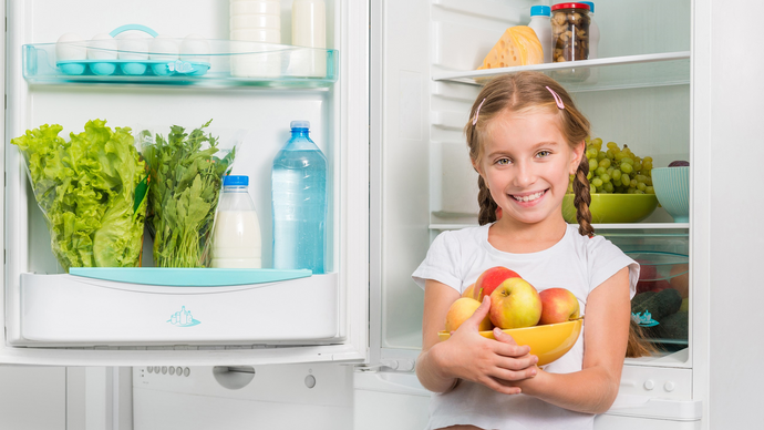 Young girl holding a bowl of fruit is standing in front of an open fridge 