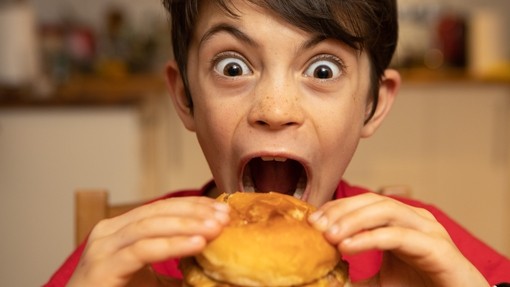 A young child makes a funny face while biting into a burger.