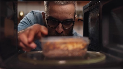 A white man wearing glasses putting a container of leftover food into a microwave.