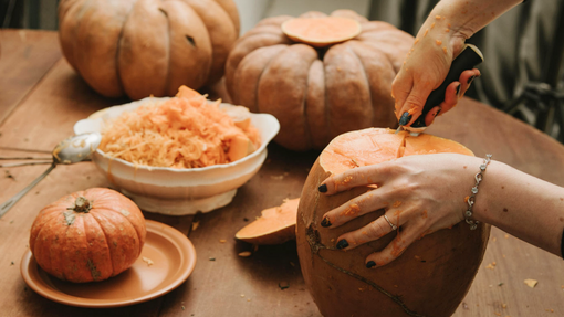 A photo of two people's hands cutting and gutting a couple of different sized orange pumpkins on a brown surface. A bowl of pumpkin flesh is also on the table.