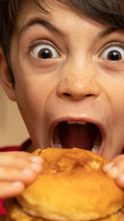 A young child makes a funny face while biting into a burger.
