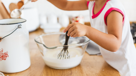 a baking bowl with white mix, a toddler is holding a whisk in the bowl and has a spoon in the other hand dipping in a pot
