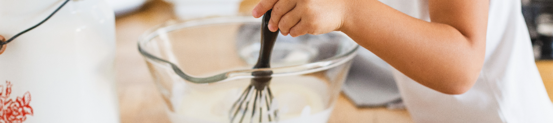 A mixing bowl with white mix and a whisk in it being held by a small child.